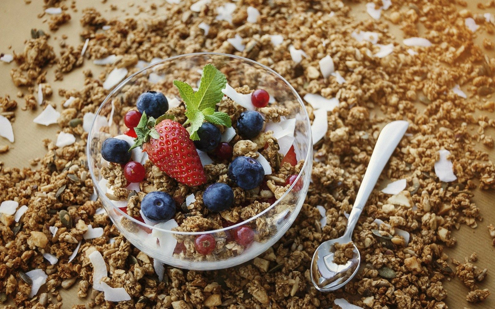 Glass bowl of granola topped with strawberries, blueberries and mint, surrounded by scattered granola