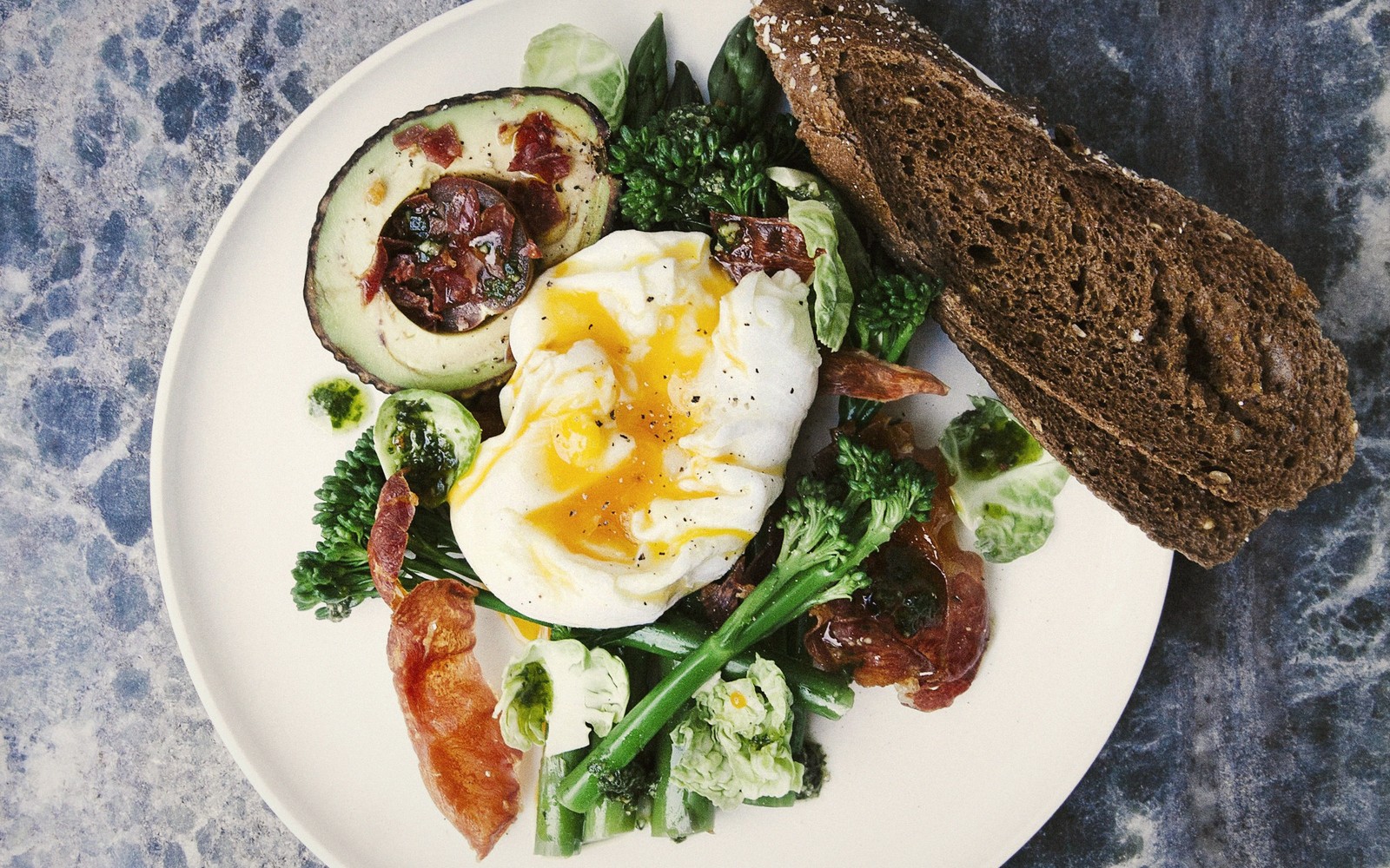 Poached egg with avocado, prosciutto, broccolini and rye bread on a plate