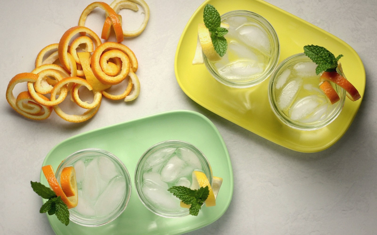 Four iced citrus drinks garnished with mint and orange peel beside curled orange rind spirals