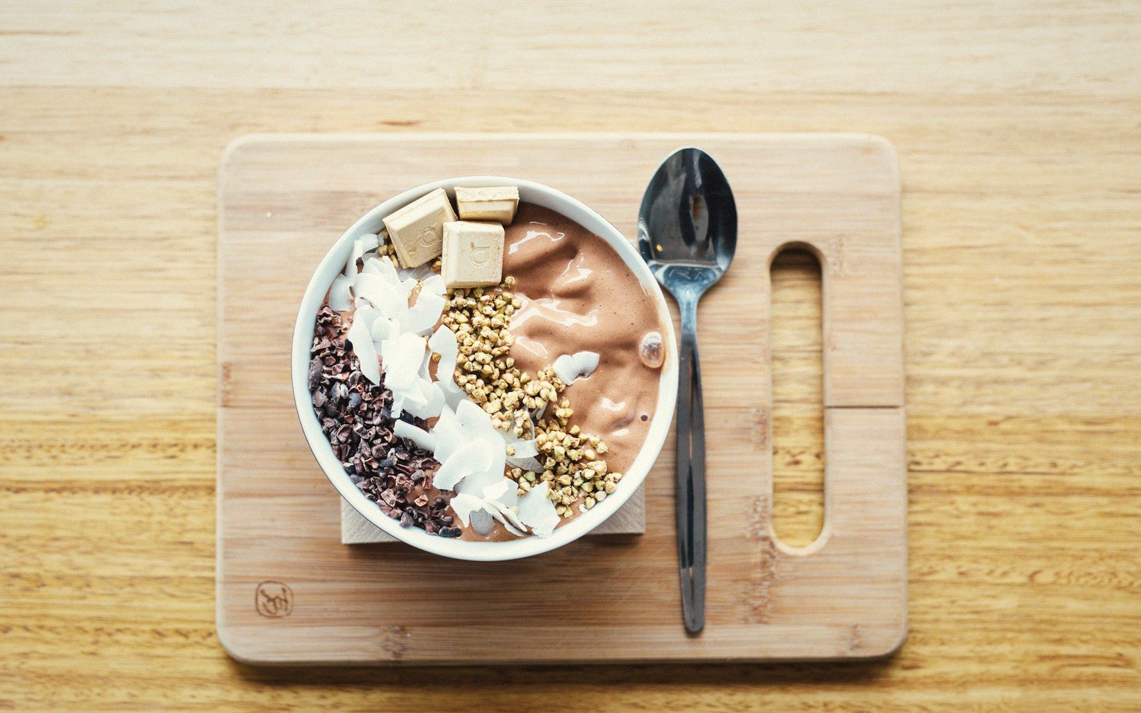 Chocolate smoothie bowl topped with cacao nibs, coconut flakes, granola and chocolate squares, with a spoon on a wooden board